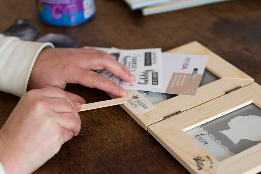 Eine Hand trägt auf einem Bilderrahmen einen Rubbelsticker der Marke Heyda auf. Mit einem Holzstäbchen wird der Sticker auf das Holz gedrückt und so direkt übertragen.