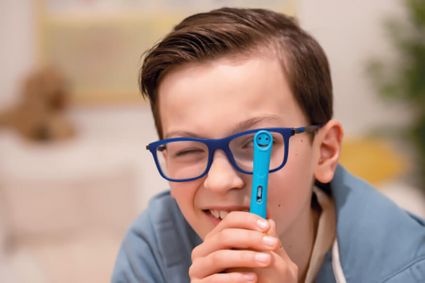 Ein Junge mit einer dunkelblauen Brille hält sich einen Griffix-Anfängerfüller von Pelikan vor das linke Auge. Das andere Auge ist zugekniffen. Im Hintergrund ist unscharf ein Kinderzimmer zu erkennen.