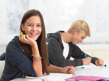 Zwei Jugendliche sitzen gemeinsam am Tisch und machen Schulaufgaben. Der Junge im Hintergrund schaut auf sein Heft und schreibt mit einem Füller. Das Mädchen im Vordergrund stütz das Kinn auf den Arm und schaut in die Kamera. In der Hand hält Sie einen Twist-Füller von Pelikan. Der Hintergrund ist hell