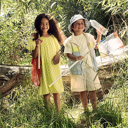 Ein Mädchen und ein Junge stehen mit Käscher und Netzen in der grünen Natur. Beide sind sommerlich gekleidet. Der Junge hält eine leere Plastikflasche hoch. Im Hintergrund sieht man einen umgestürzten Baum.
