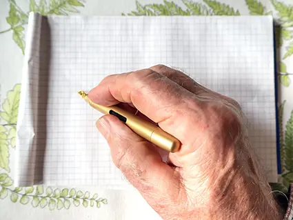 Eine alte, männliche Hand schreibt mit einem goldenen Füller von Kaweco auf einen karierten Block. Der Untergrund ist eine Tischdecke mit grünem Farn-Muster. Foto: Johanna Rundel 