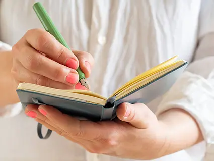 Eine Frau in weißer Bluse schreibt mit einem hellgrünen Füller von Kaweco in ein aufgeschlagenes, blaues Notizbuch, dass sie in ihren Händen hält. Foto: Johanna Rundel 