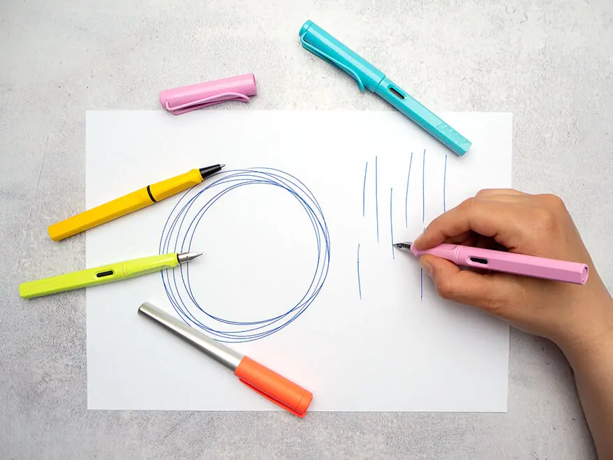 Jemand malt mit einem rosa Füller von Lamy große Kreise und Striche auf ein weißes Blatt Papier. Weitere, bunte Schreibgeräte sind darum verteilt.