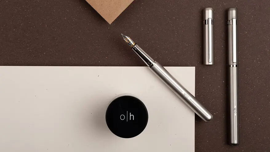 Auf einem dunkelbraunen Untergrund liegt ein weißes Blatt Papier. Darauf steht ein Tintenfass sowie zwei silberne Füller.