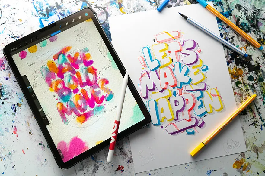 Ein weißes DIN A4 Blatt mit einem bunten Buchstaben-Lettering liegt auf einem vollgeklecksten Untergrund. Daneben ein Tablet mit einem digitalen Lettering. Drumherum liegen verschiedene Stifte von Stabilo.