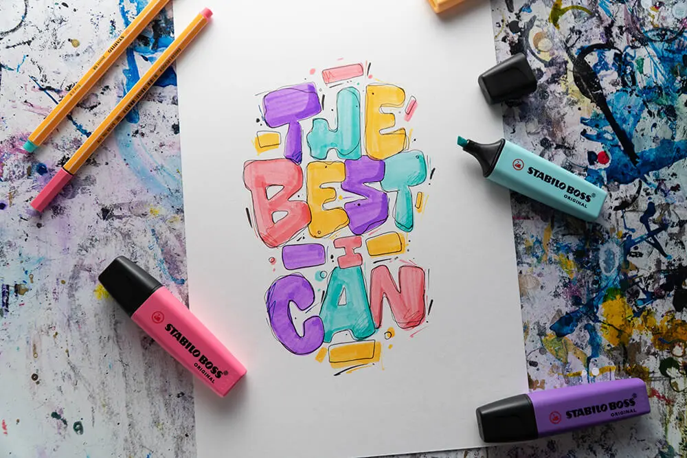 Ein weißes DIN A4 Blatt mit einem bunten Buchstaben-Lettering liegt auf einem vollgeklecksten Untergrund. Drumherum liegen verschiedene Stifte von Stabilo.