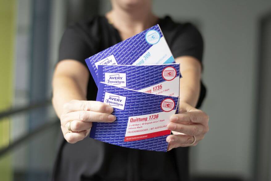 Eine Frau hält drei verschiedene Quittungsblöcke von Avery Zweckform aufgefächert in die Kamera.