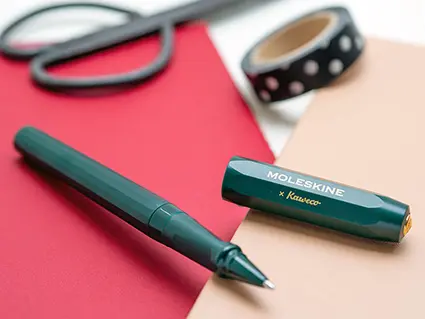 Ein dunkelgrüner Kugelschreiber mit der Aufschrift Moleskine liegt auf zwei Notizbüchern. Im Hintergrund ist eine Rolle Washitape und eine Schere zu sehen. Foto: Johanna Rundel