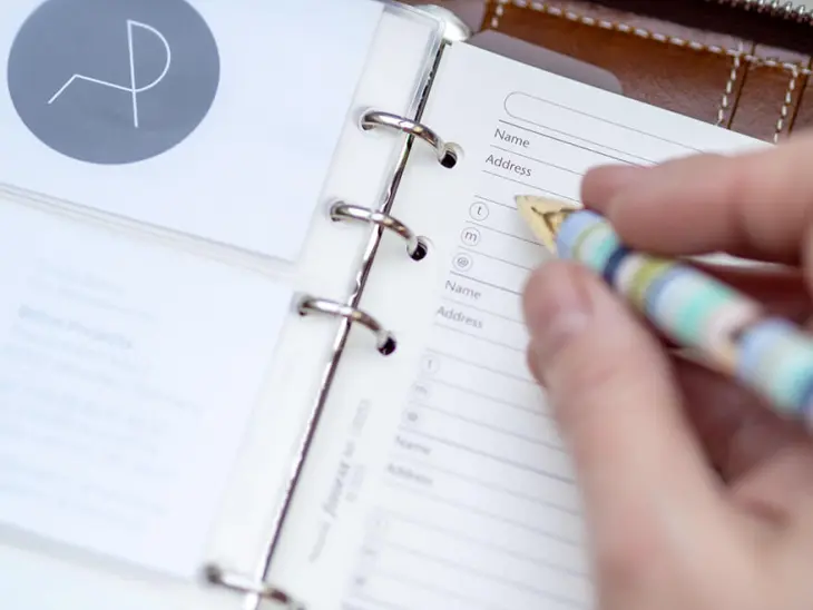 Jemand schriebt mit einem pastellfarbenen Kugelschreiber in einen braunen Lederplaner von Filofax. Daneben ist ein Einhefter für Visitenkarten. Foto: Johanna Rundel 