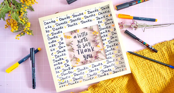 Auf einem rosa Untergrund liegt ein selbstgestalteter Bilderrahmen mit einem Lettering. Darum liegen verschieden Marker von POSCA und Faber-Castell. Foto: Johanna Rundel