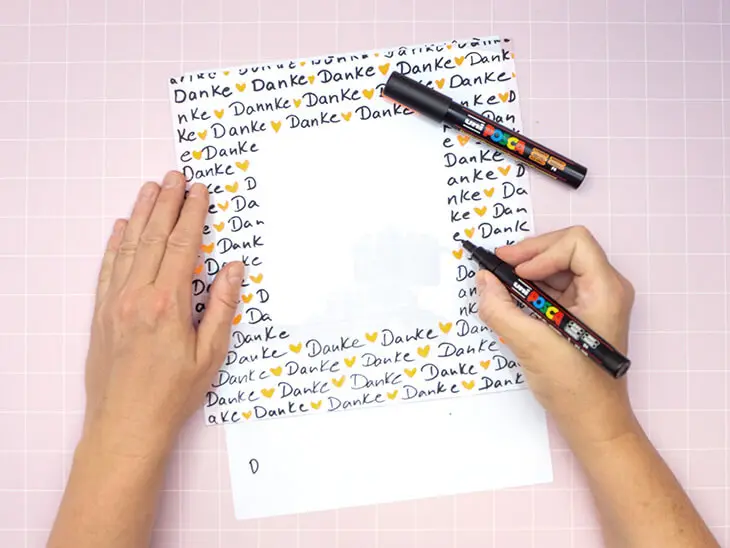 Jemand schreibt mit einem schwarzen Marker von POSCA ganz oft das Wort „Danke“ auf das Glas eines quadratischen Bilderrahmens. Foto: Johanna Rundel