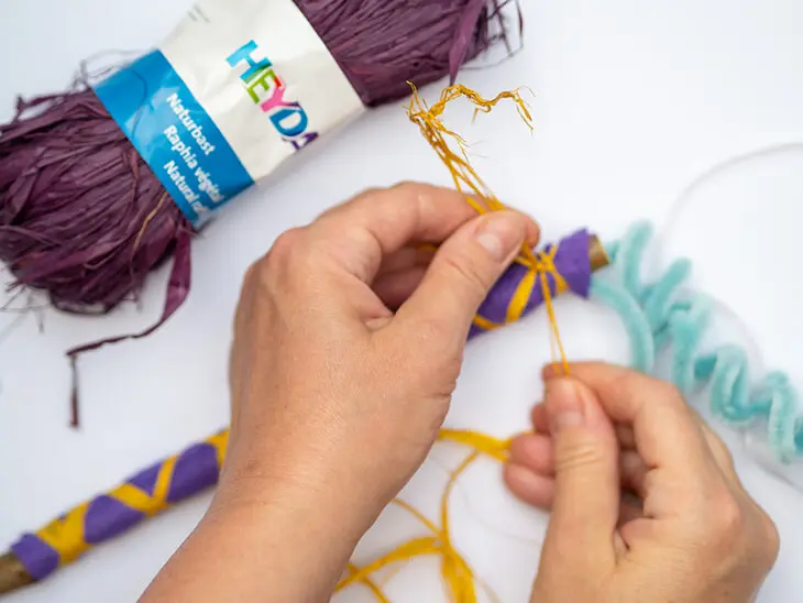 Jemand umwickelt einen Stock, der mit violettem Bastelfilz von KNORR prandell beklebt wurde mit gelbem Naturbast von HEYDA. Foto: Johanna Rundel