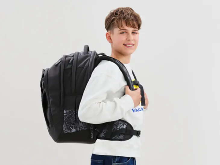 Ein Jugendlicher mit einem dunkeln Schulrucksack von satch steht vor einem weißen Hintergrund.