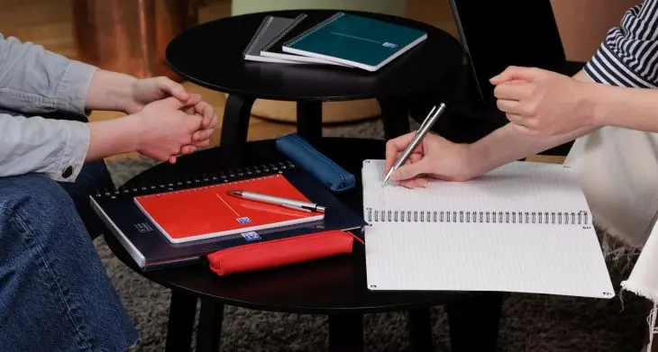 Auf einem niedrigen Tisch liegen Notizblöcke von Oxford, in die Hände etwas hineinschreiben