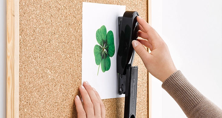Auf dem Bild befestigt eine Person ein Blatt Papier mit einem grünen Kleeblatt-Motiv auf einer Korktafel. Dabei wird ein Novus-Heftgerät verwendet, um das Papier sicher an der Pinnwand zu fixieren.