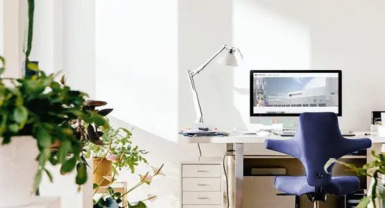 Blick auf einen hellen Homeofficeplatz mit Schreibtisch, Bildschirm, Bürostuhl, Lampe und Pflanzen.