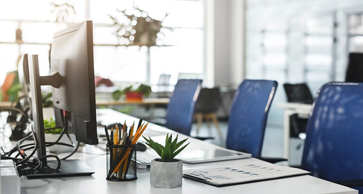 Blick in ein Büro, an dem drei Arbeitsplätze nebeneinander an einem Tisch installiert sind.