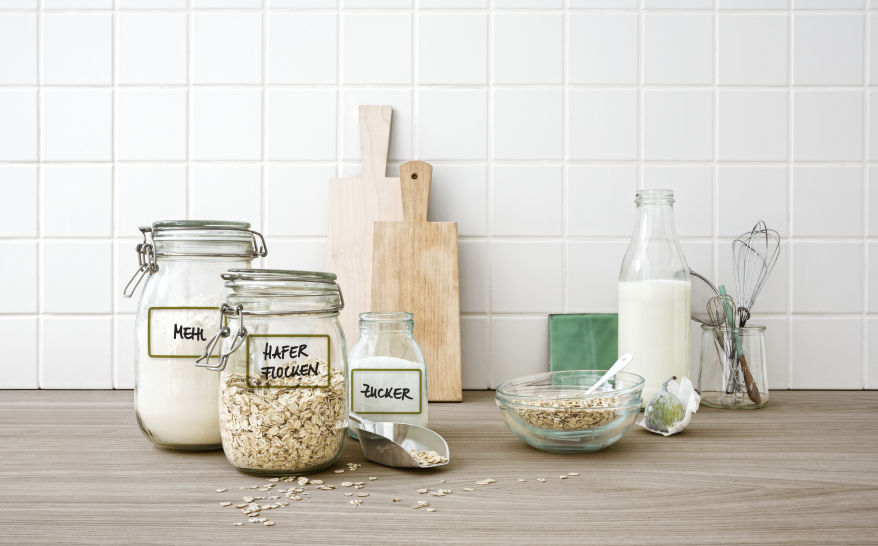 Auf einer Küchenanrichte stehen diverse Einmachgläser, welche mit Tafelaufklebern der Marke Avery Zweckform beklebt und mit "Mehl", "Haferflocken", "Zucker" beschriftet sind. An der Wand lehnen 3 Brettchen, es steht eine Glasfalsche mit Milch auf der Anrichte und eine Glasschüssel mit Haferflocken. Ein Glas mit 2 Schneebesen stehen außerdem auf der Anrichte.