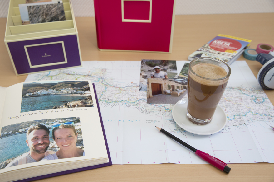 Auf einem Tisch liegen ein aufgeschlagenenes Fotoalbum mit Urlaubsbilder, ein weiteres Fotoalbum eund eine Fotobox der Marke Semikolon liegen auf dem Tisch. Eine Landkarte ist ausgebreitet, darauf liegen Urlaubsfotos und es steht ein Latte Macchiato auf der Karte.