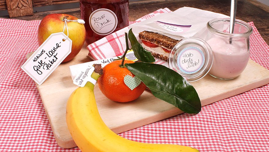 Auf einer rot-weiß karierten Tischdecke liegt ein Holzbrettchen. Darauf liegen eine Banane, eine Mandarine, ein Apfel, der ein Etikett der Marke Avery Zweckform angehängt hat mit der Aufschrift "Kleines Gute-Laune Paket". Auf dem Brettchen steht außerdem ein kleines Weckglas, welches einen Etiketten-Aufkleber mit der Aufschrift "Hab dich lieb!" Eine Butterbrot-Tüte mit einem Brot ist auch mit einem Etikett beklebt.