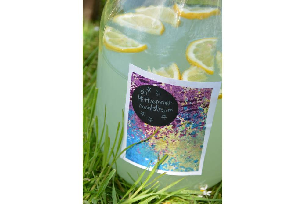 Nahaufnahme einer großen Wasserflasche, die im Gras steht. Auf der Flasche ist ein selbst gestaltetes buntes Etikett mit der Aufschrift "Mittsommernachts-Traum".