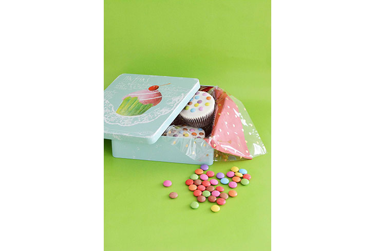 In einer Metalldose mit dem Motiv eines Cupcakes auf dem Deckel sind Cupcakes zu sehen. Die Dose ist mit Servietten ausgelegt und vor der Dose liegen bunte Smarties.