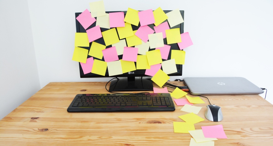 Auf einen Tisch steht ein Computer-Monitor, der mit Post-its beklebt ist. Davor liegt eine Tastatur, daneben liegt ein angeschlossener Laptop.