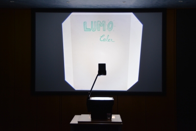 In einem dunklen Raum steht ein Overhead-Projektor, welcher eine Folie mit dem Wort "Lmocolor" an die Wand projiziert.