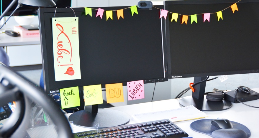 An einem Monitor im Büro hängen eine Papiergirlande und Post-its mit Sprüchen zum Geburtstag.