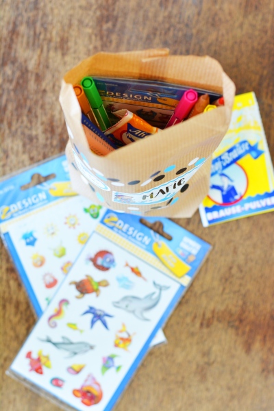 Auf einem Holzboden steht eine geöffnete Papiertüte, die mit Stickern und Etiketten verziert wurde. Diese ist mit Stickern von Avery Zweckform, Stiften und Brause gefüllt. Die Tüte steht auf gefüllten Packungen mit Stickern von Avery Zweckform und einer Packung Brause.