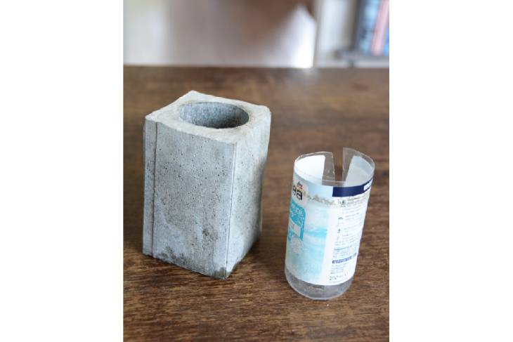Auf einem Holztisch steht der ausgehärtete Betonbescher. Daneben steht die Plastikflsche, welche aus dem Betonbecher entfernt wurde.