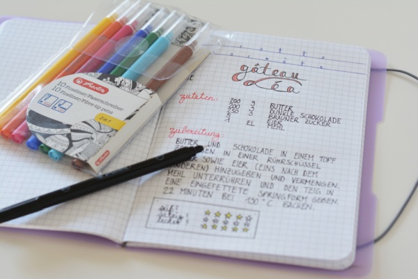 Auf einem weißen Tisch liegt ein aufgeschlagenes my.book flex von Herlitz mit lilafarbenen Einband. Darin wurde ein Rezept für einen Schokoladenkuchen notiert. Auf dem Notizbuch liegen ein Set Fasermaler der Marke Herlitz. Ein Marker liegt geöffnet daneben.