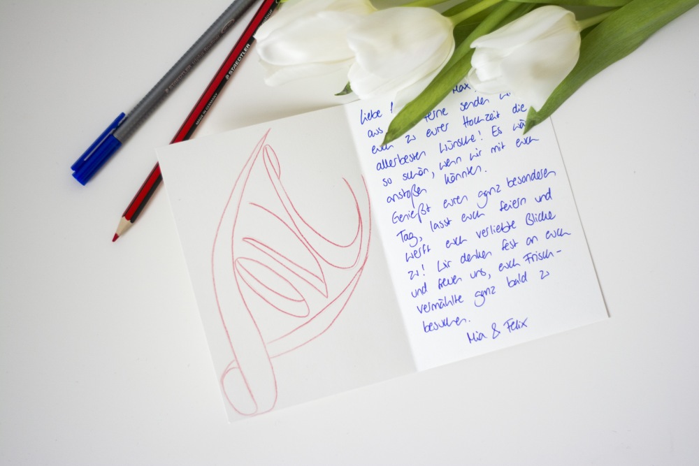 Auf einem weißen Tisch liegt eine aufgeklappte, selbst gestaltete Hochzeitskarte. Auf der linken Seite ist mit rotem Buntstift der Schriftzug "LoveW gelettert, auf der rechten Seite steht der Text der Karte. Es leigen weiße Tulpen, ein blauer Fineliner und ein roter Buntstift der Marke Staedtler auf dem Tisch.
