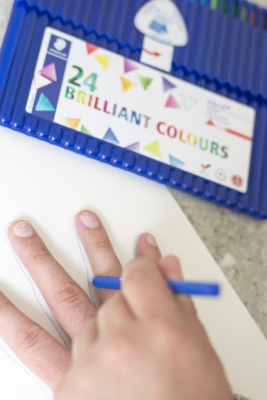 Hand wird mit Buntstift auf Papier nachgezeichnet, Verpackung von Farbstifte daneben