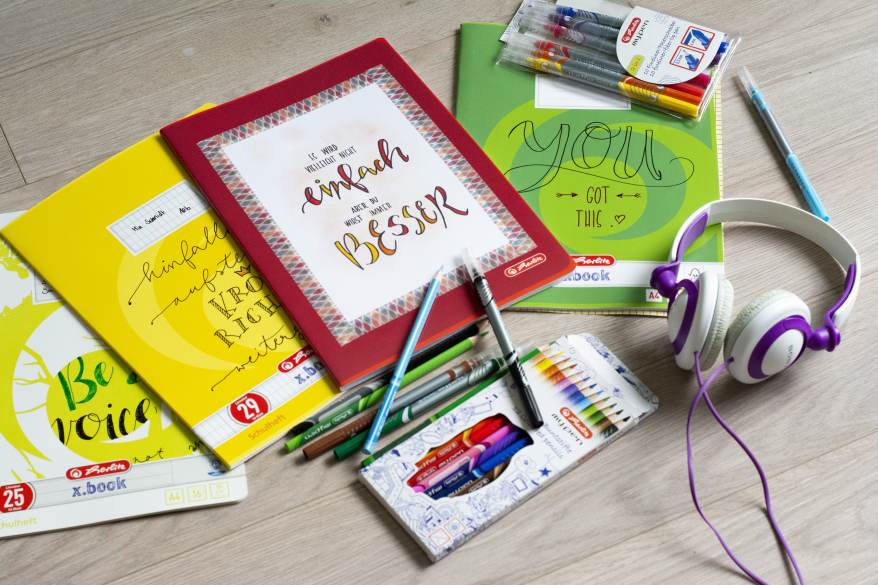 Auf einem Tisch liegen verschieden beletterte Schulhefte sowie Filz- und Buntstifte der Marke Herlitz. Außerdem liegt ein Kopfhörer daneben.