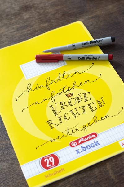 Ein gelbes Schulheft der Marke Herlitz ist mit dem Spruch "Hinfallen, aufstehen, Krone richten, weitergehen" belettert. Auf dem Heft liegen 2 Colli Marker von herlitz.