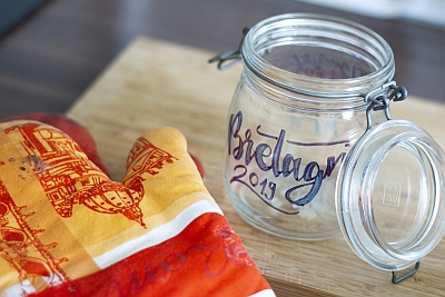 Ein Weckglas mit dem in blau geletterten Schriftzug "Bretagne 2019" steht auf einem Tisch. Daneben liegt ein Ofenhandschuh in rot und gleb.