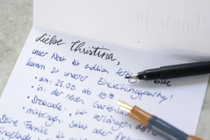 Eine aufgeklappte Einladungskarte, auf der die Anrede gelettert ist. Der restliche Text wurde mit Füller geschrieben, der auf der Karte liegt.