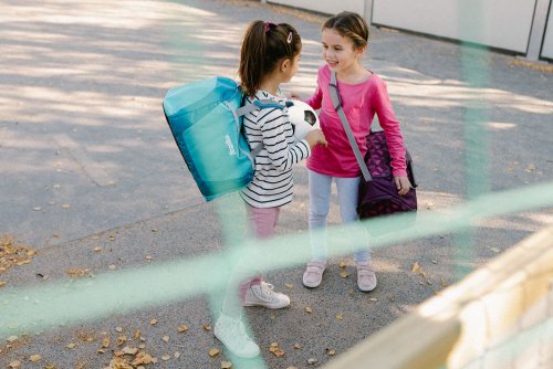 Zwei Mädchen im Grundschul-Alter stehen auf dem Schulhof. Beide haben eine Sporttasche der Marke ergobag umhängen.