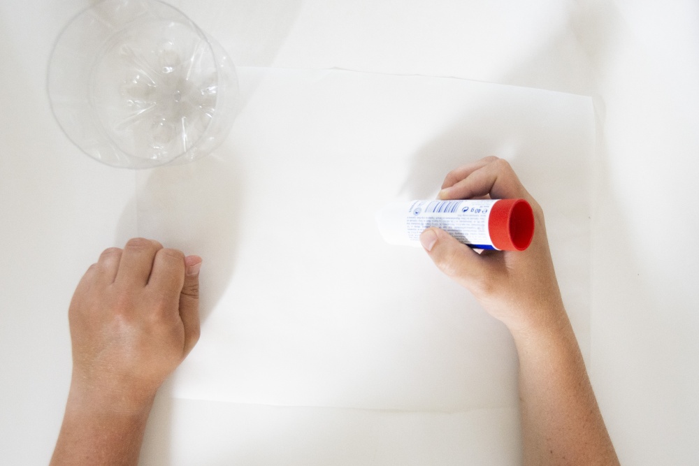 Hände tragen Kleber auf ein weißes Papier auf, daneben liegt eine abgeschnittene Plastikflasche.