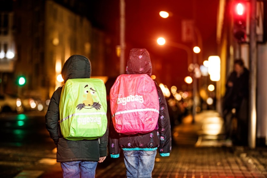 Zwei Kinder in Regenkleidung tragen zwei Schulranzen, die mit Regencapes in Neonfarben geschützt sind.
