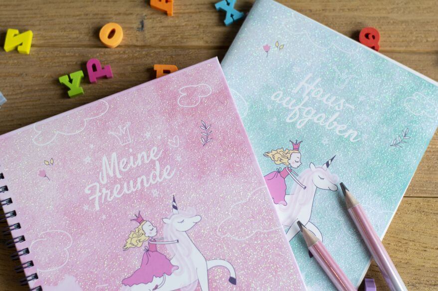 Zwei Hefte in Einhorndesign mit den Aufschriften "Meine Freunde" und "Hausaufgaben" liegen auf einem Schreibtisch. Darum sind zwei Bleistifte und Radiergummis in ABC-Buchstabenform platziert.