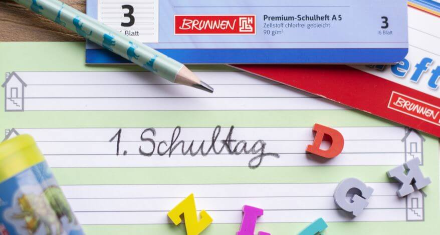 In ein Schreiblernheft der 1. Grundschulklasse steht in Schreibschrift "1. Schultag" geschrieben. Darum liegen zwei Hefte, ein Bleistift, ein Anspitzer und Radiergummis in ABC-Buchstabenform.
