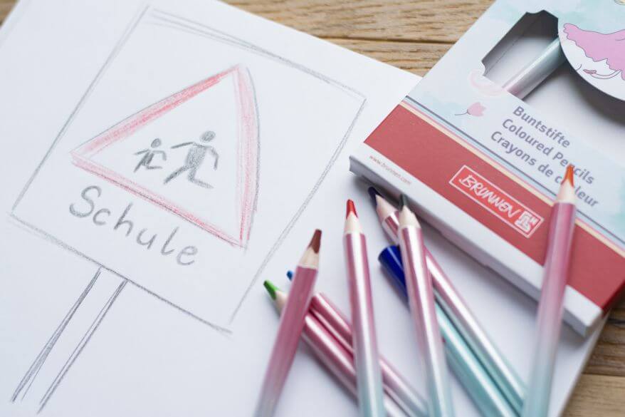 Auf ein Blatt Papier ist ein Verkehrsschild "Schule" aufgemalt. Daneben liegen Buntstifte von Brunnen.