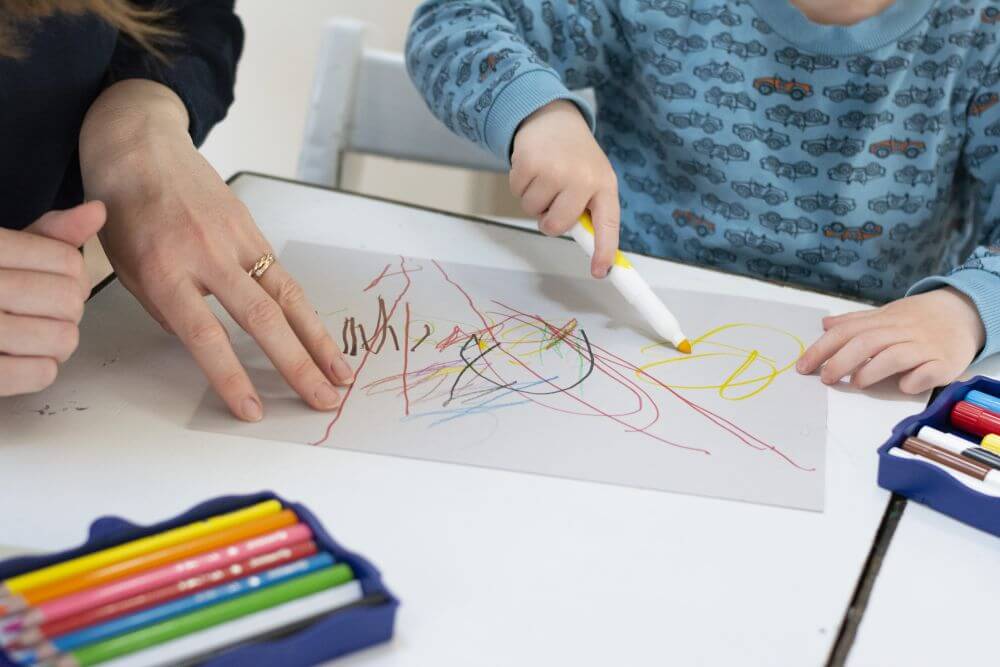 Eine Kinderhand greift nach Filzstiften aus der Kreativfabrik von Pelikan und malt Muster auf ein Blatt Papier, daneben hält die Hand eines Erwachsenen das Papier fest.