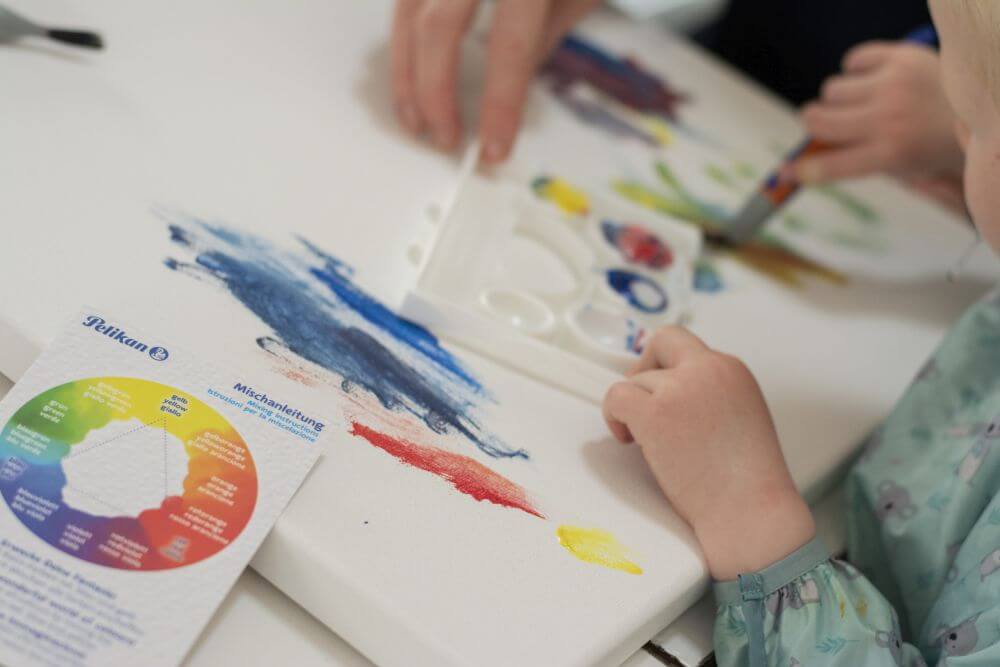 Eine Kinderhand malt mit Pinsel und Gouache aus der Kreativfabrik von Pelikan auf eine Leinwand, im Vordergrund liegt eine Mischanleitung für Farben.