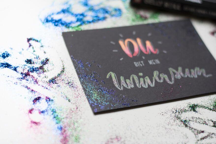 Nahaufnahme des Handletteringschriftzugs "Du bist mein Universum", der mit Gelstiften "Gelly Rolls" von Sakura gestaltet wurde. Das Wort "Universum" glitzert, drumherum liegt Glitter verstreut.