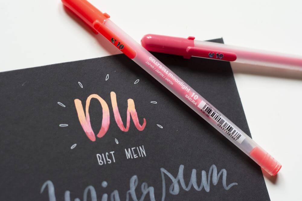 Nahaufnahme eines selbstgemachten Handletterings "Du bist mein Universum, das mit Gelstiften "Gelly Rolls" von Sakura gestaltet wurde. Das Wort "Du" leuchtet in fluoreszierenden Farben der Gelstifte "moonlight".