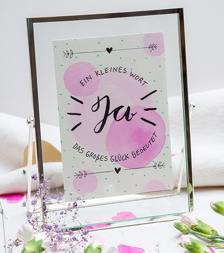 Vor einem hellen Hintergrund steht ein silberner Bilderrahmen mit Aufsteller von Goldbuch. Er besteht aus zwei aufeinanderliegenden Glasscheiben, zwischen denen eine Aquarellkarte klemmt. Auf der Karte steht: „Ja – ein kleines Wort, das großes Glück bedeutet.“. Der Spruch ist auf große, rosa Aquarellpunkte gelettert. Um den Rahmen liegen pinke und rosa Herzen aus Papier und verschiedene Blumen verteilt. Foto: Johanna Rundel