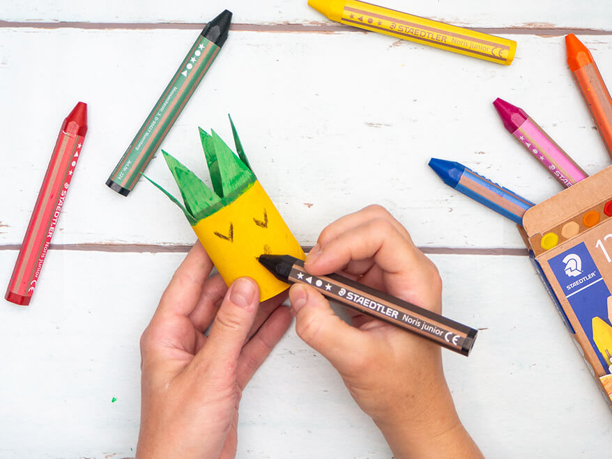 Jemand malt mit einem schwarzen Buntstift von Staedtler "V's" auf den gelben Teil einer zur Ananas umgestalteten, leeren Klopapierrolle. Drumherum liegen Buntstifte in weiteren Farben. Der Untergrund ist hell. Foto: Johanna Rundel